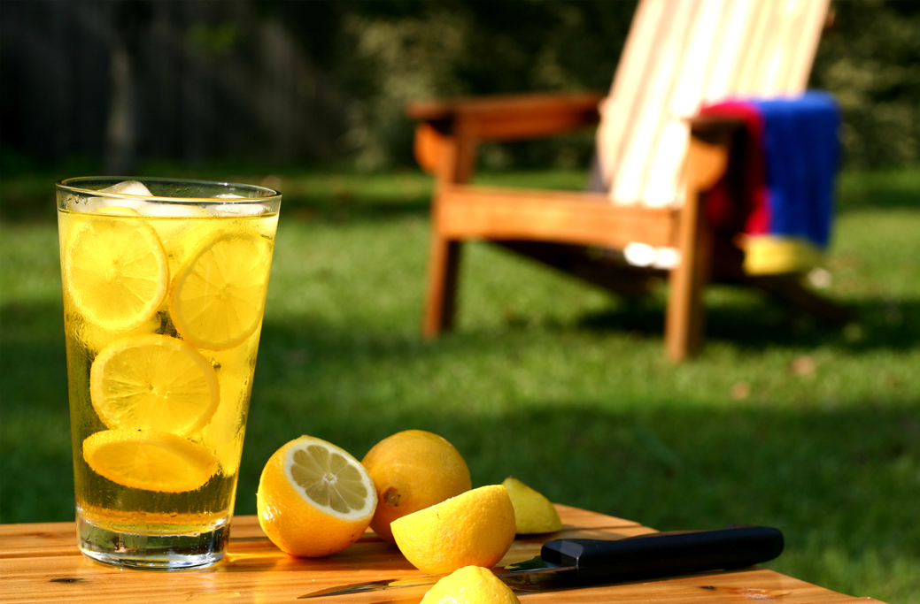 Beverage with lemon slices in it on a table in a freshly landscaped lawn. Some cut lemons sitting on table next to glass. Paring knife beside lemons on table.