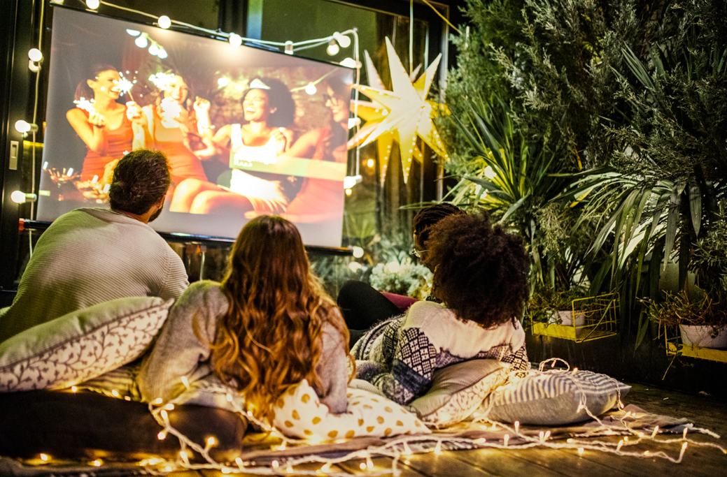 Three people enjoying movie night on a backyard movie projector with string lights and plants.